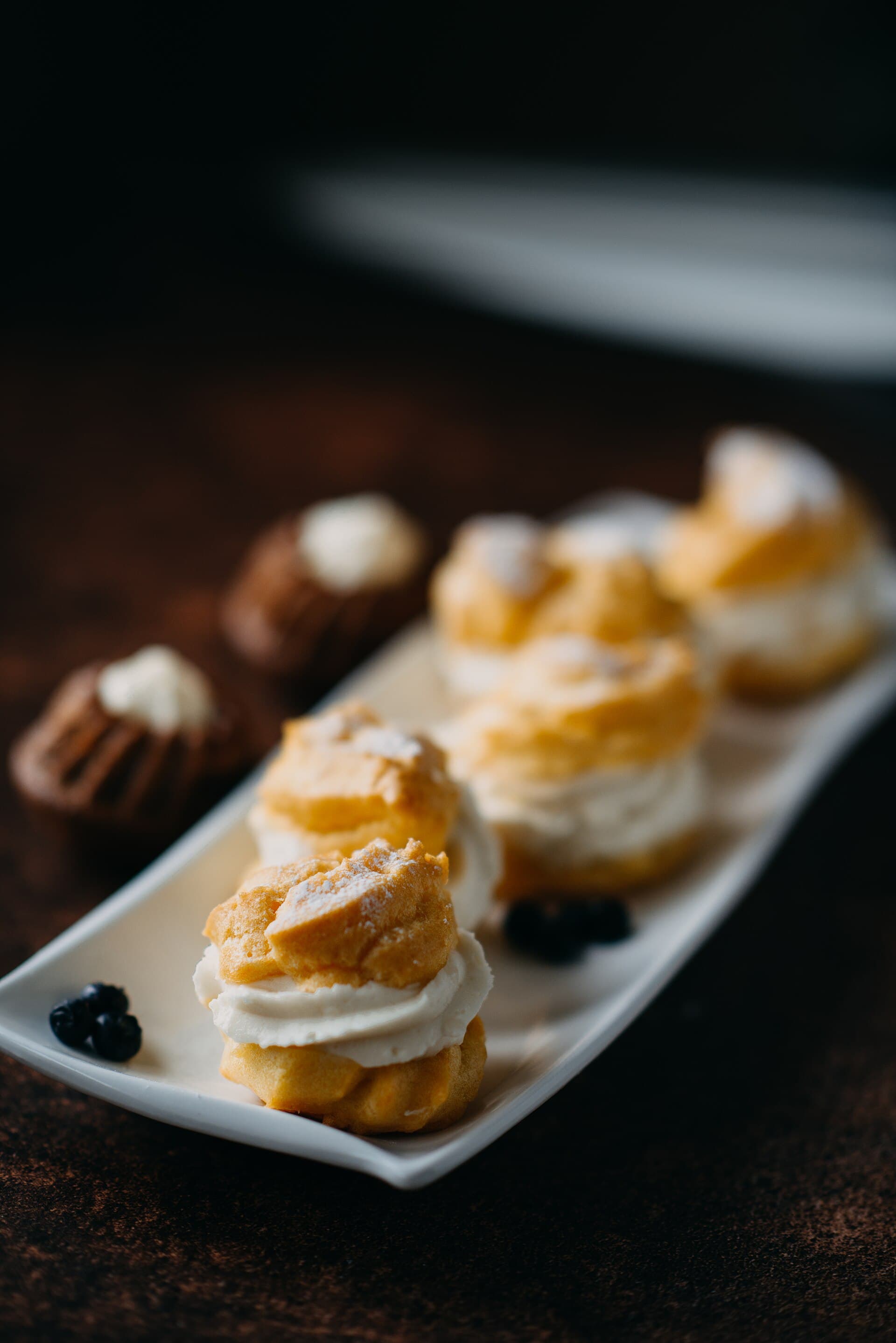 Choux rellenos de crema y pequeños pasteles de chocolate en una bandeja blanca, sobre fondo oscuro