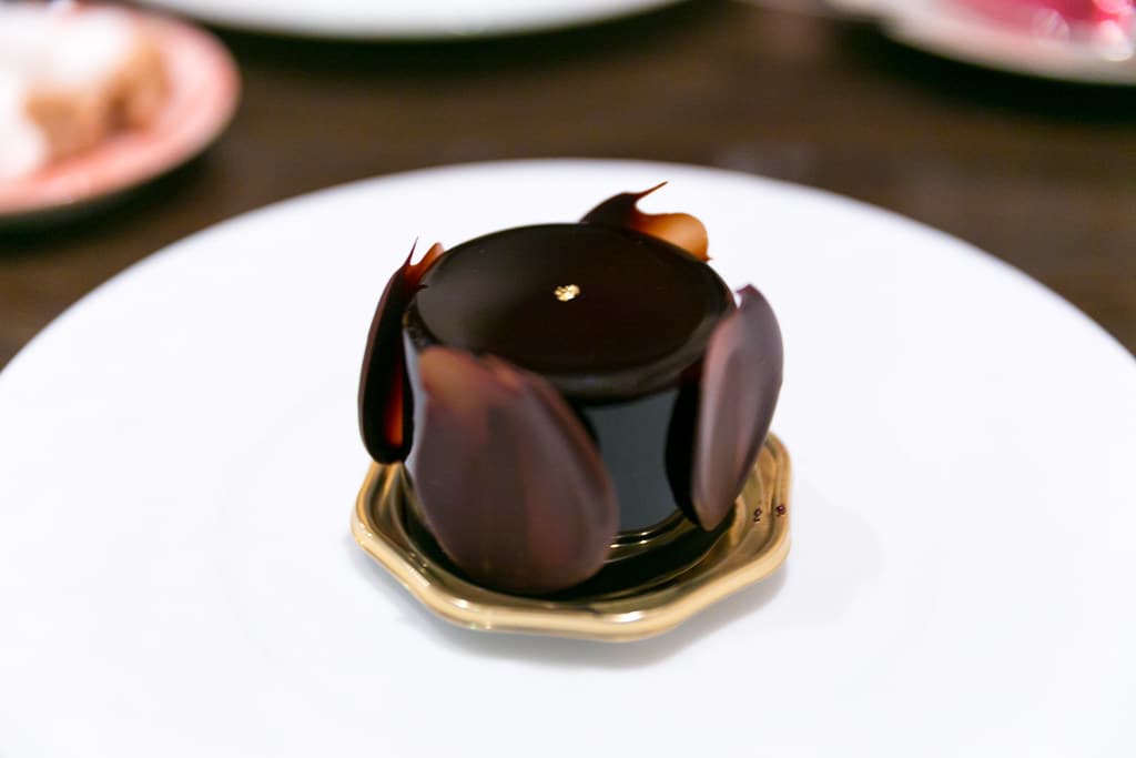 Entremet de chocolate rodeado de pétalos de chocolate, sobre un plato blanco